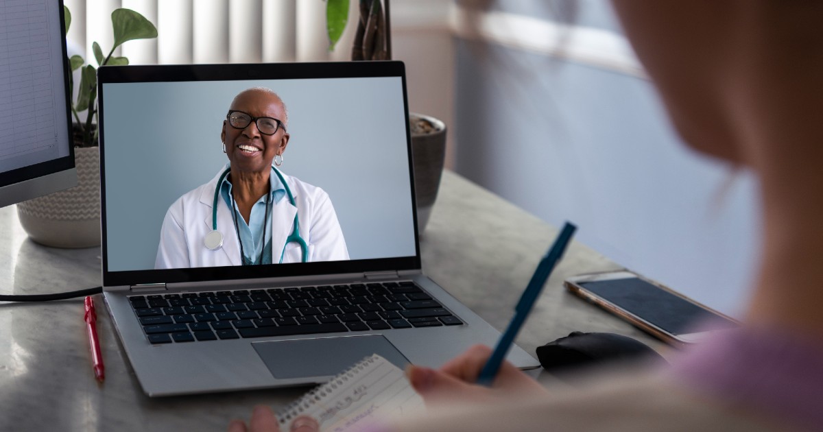 Female doctor on laptop smiles at patient in foreground who is taking notes Female doctor on laptop smiles at patient in foreground who is taking notes
