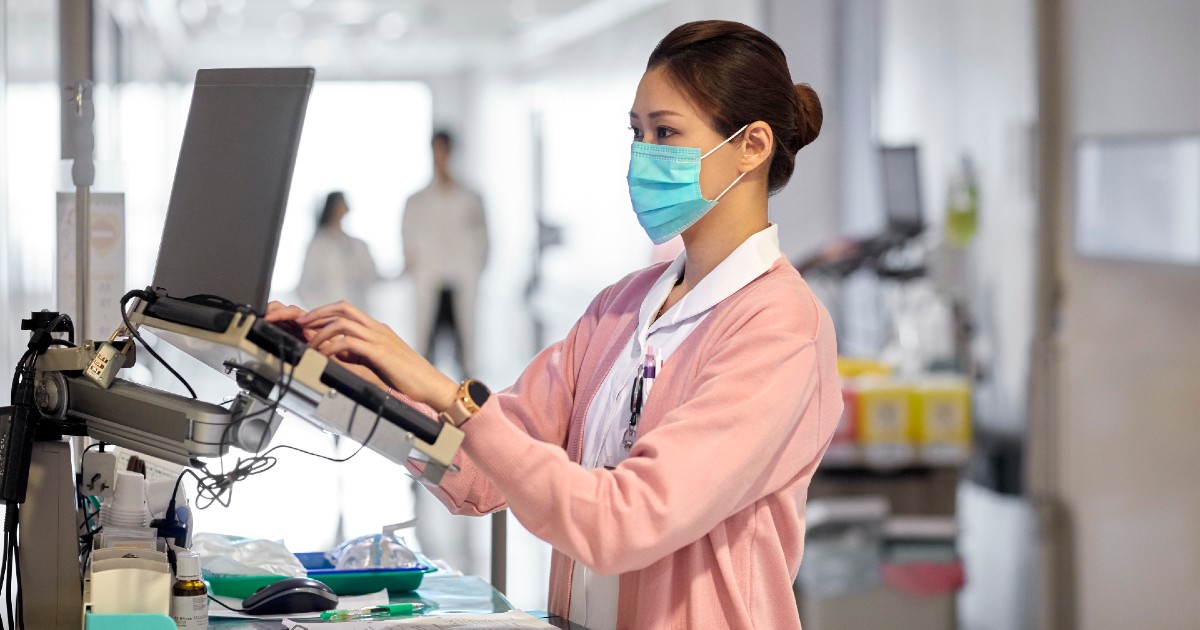 Nurse in mask and pink sweater types notes in a hospital hallway Nurse in mask and pink sweater types notes in a hospital hallway